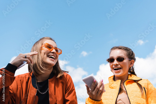 Fotografía woman in stylish sunglasses holding smartphone near happy friend against blue sky