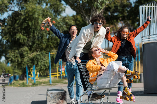 young and cheerful multiethnic skaters having fun with shopping cart in park.