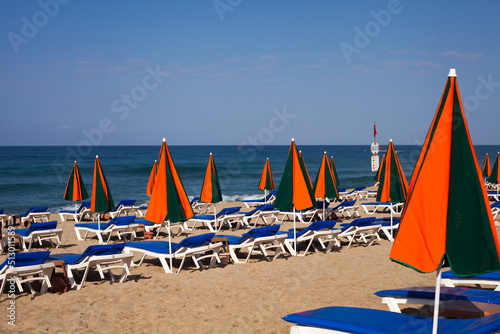 Beach with closed striped umbrellas and deck chairs on the background of the sea. An equipped beach is waiting for guests to relax. Orange and green stripes. Mediterranean Sea