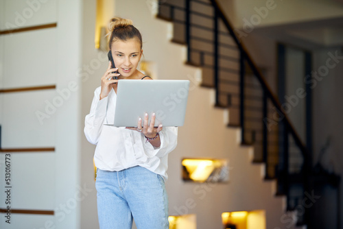 A woman at home with a laptop in hand is having a phone call and smilling. Stairs on the background. Indoor