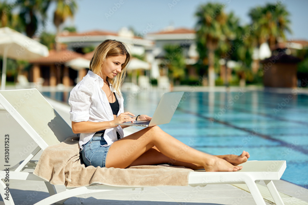 A beautiful woman is working on the laptop sitting on the sun lounger near the pool. Outdoor