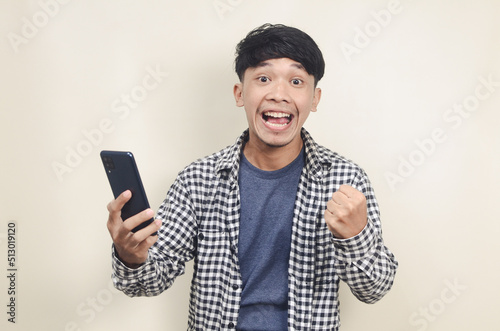 Close-up portrait of happy young Asian male model wearing plaid shirt with winning expression while holding phone