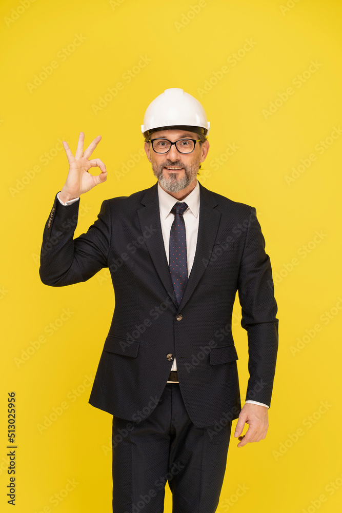 Gesturing OK handsome middle aged businessman, architect in construction helmet looking at camera isolated on yellow background. Handsome mature head of construction in black suit. Architect concept. Gesturing OK handsome middle aged businessman, architect in construction helmet looking at camera isolated on yellow background. Handsome mature head of construction in black suit. Architect concept.