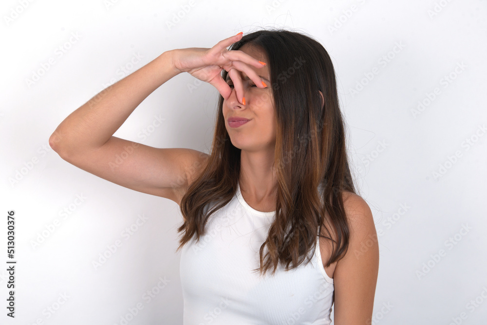 Displeased young beautiful caucasian woman wearing white Top over white background plugs nose as smells something stink and unpleasant, feels aversion, hates disgusting scent. Displeased young beautiful caucasian woman wearing white Top over white background plugs nose as smells something stink and unpleasant, feels aversion, hates disgusting scent.