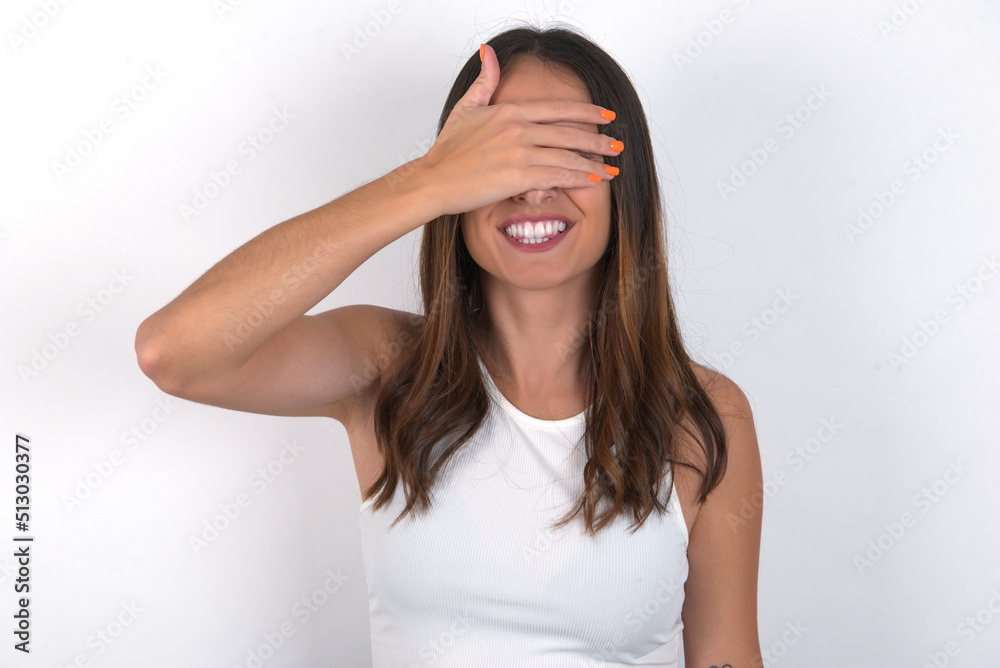 Happy young beautiful caucasian woman wearing white Top over white wall closing eyes with hand going to see surprise prepared by friend standing and smiling in anticipation for something wonderful. Happy young beautiful caucasian woman wearing white Top over white wall closing eyes with hand going to see surprise prepared by friend standing and smiling in anticipation for something wonderful.