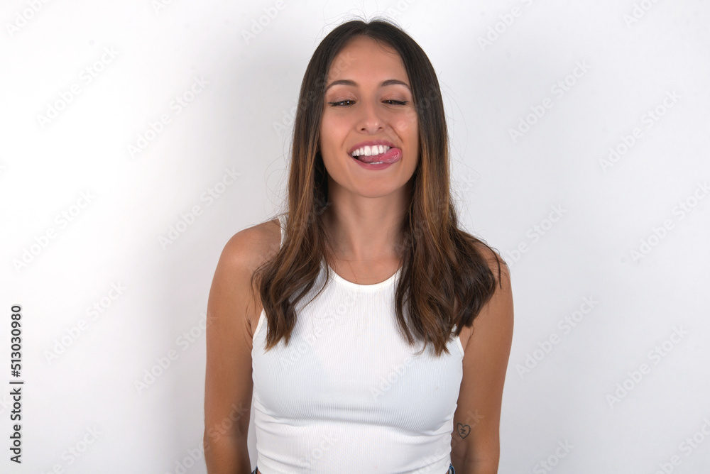 Funny young beautiful caucasian woman wearing white Top over white background makes grimace and crosses eyes plays fool has fun alone sticks out tongue. Funny young beautiful caucasian woman wearing white Top over white background makes grimace and crosses eyes plays fool has fun alone sticks out tongue.