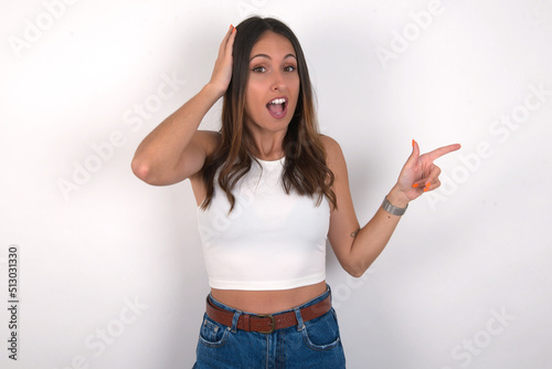 Surprised young beautiful caucasian woman wearing white Top over white background, pointing at empty space holding hand on head