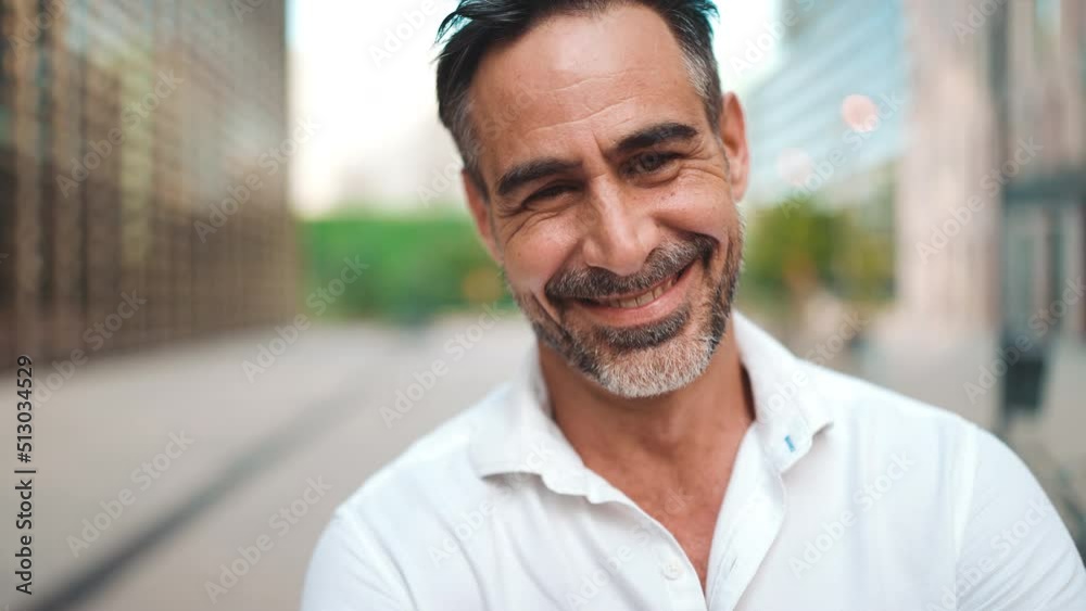 Close-up mature businessman with neat beard is looking at the camera in the financial district in the city. Successful man smiling at the camera on modern buildings background Close-up mature businessman with neat beard is looking at the camera in the financial district in the city. Successful man smiling at the camera on modern buildings background