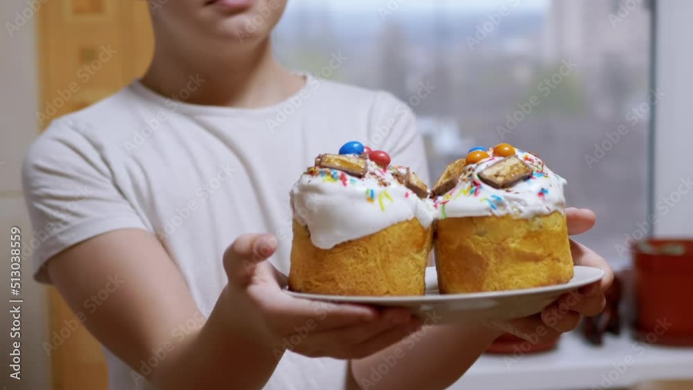 Boy, Standing in the Kitchen, Holds Out Two Easter Cakes on a Plate. Close-up. Yummy prepared Easter cupcakes decorated with white icing, and multicolor sprinkles. Orthodox holiday of Easter. Zoom. Boy, Standing in the Kitchen, Holds Out Two Easter Cakes on a Plate. Close-up. Yummy prepared Easter cupcakes decorated with white icing, and multicolor sprinkles. Orthodox holiday of Easter. Zoom.