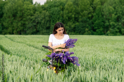 A pretty girl stands with a bouquet of lupines in a field next to a bicycle © Julia Jones