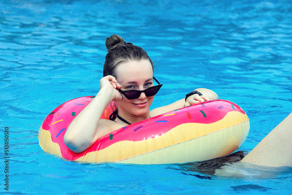 portrait of a beautiful girl in sunglasses, she has fun in the water of an outdoor pool in the summer, she is wearing a bright red rubber ring