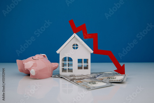 Wooden house and a red arrow down with broken piggy bank on money banknotes. The concept of inflation and economic recession