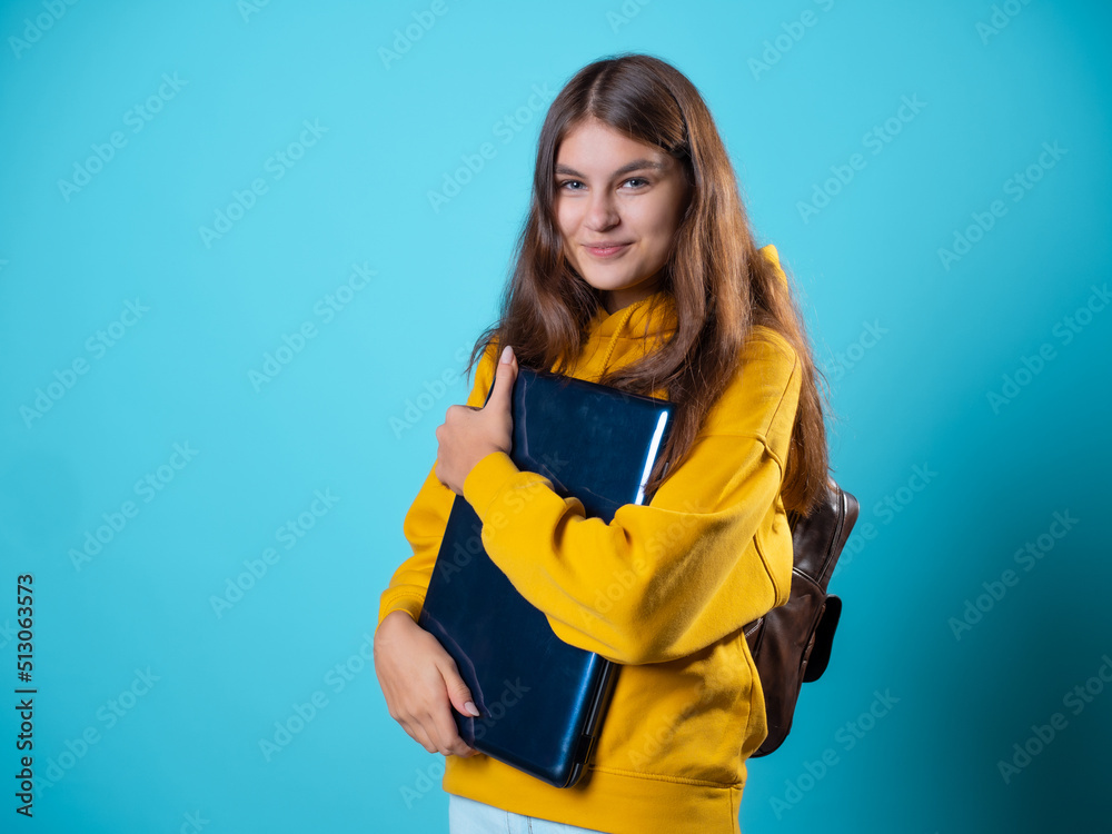Work and study remotely, a new normal, a concept. A student with a laptop in her hands, a young brunette in a yellow hoodie on a blue background Work and study remotely, a new normal, a concept. A student with a laptop in her hands, a young brunette in a yellow hoodie on a blue background