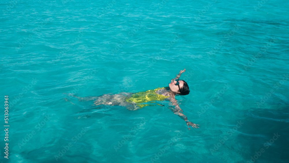 young woman in a one-piece yellow swimsuit floats on the surface of the water. Brunette girl relaxes and bathes in the blue sea, relaxing by the sea