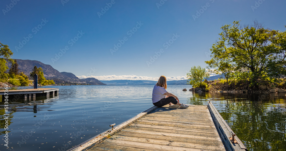 Woman relaxes by the lake sitting on the edge of a wooden jetty. Sunny summer morning or evening concept. Woman relaxes by the lake sitting on the edge of a wooden jetty. Sunny summer morning or evening concept.