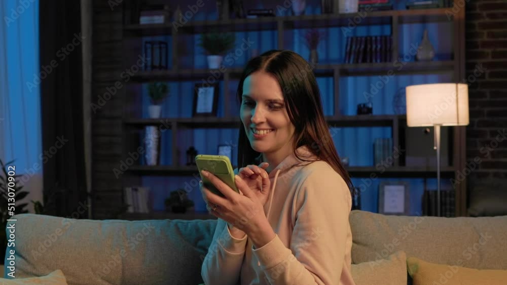 Young beautiful woman using mobile phone apps, browsing internet, social media, looking at smartphone and smiling. Home leisure in the evening, cinematic lighting. Relaxation after hard day at work. Young beautiful woman using mobile phone apps, browsing internet, social media, looking at smartphone and smiling. Home leisure in the evening, cinematic lighting. Relaxation after hard day at work.
