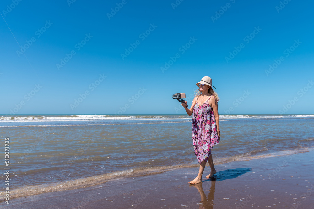 Woman in summer dress walking along the beach taking photos with a smart phone attached to a gimbal stabilizer