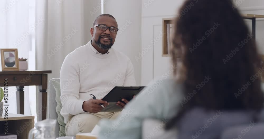 African male psychologist writing notes while listening to a female patient talk about issues and emotions during a therapy session. Young patient talking to her therapist about her problems.