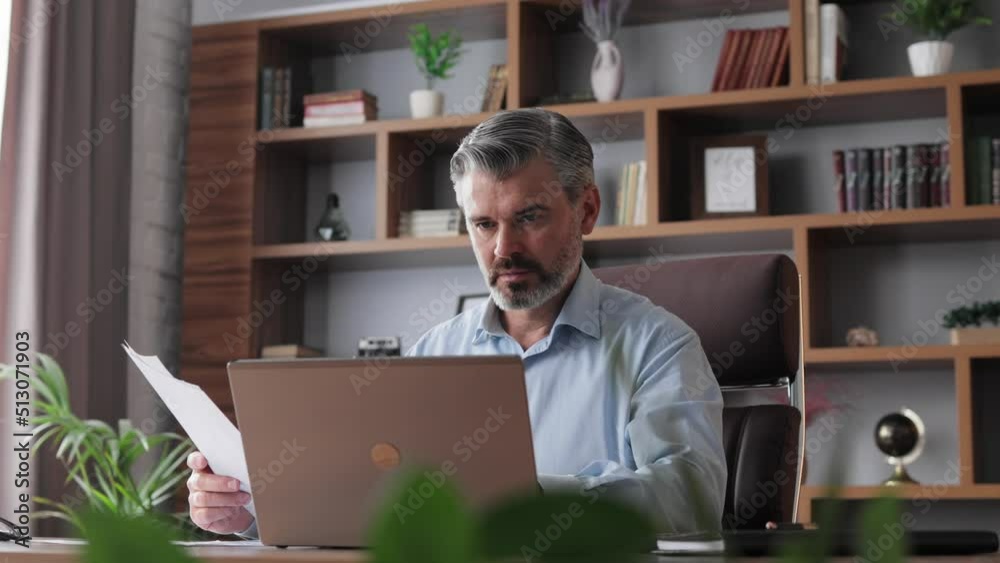 Portrait of adult gray-haired bearded businessman working with laptop at home office. Texting messages, doing paperwork, calculating monthly expenses. Business concept.