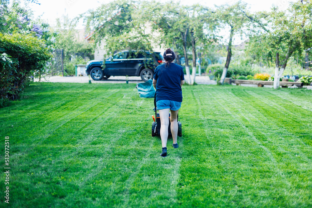 Rearview of dark-haired woman in casual clothes using lawn mower or grass cutter in the yard, ground. Cutting fresh lush green grass in countryside. Trees on background. Technological machine for lawn