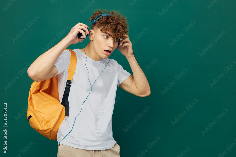 horizontal portrait of a handsome man with curly hair, standing on a green background in a gray T-shirt, and looking in surprise to the side, holding his headphones on his head with his hand horizontal portrait of a handsome man with curly hair, standing on a green background in a gray T-shirt, and looking in surprise to the side, holding his headphones on his head with his hand