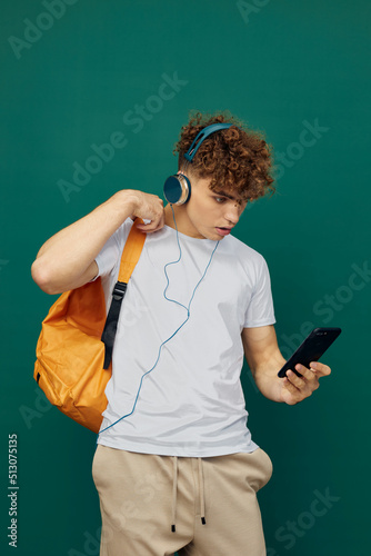 a joyful man, a teenager, stands on a green background listening to music with headphones, holding his smartphone in his hand and looking at it