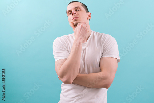 A young man holds a chin and looks down from the top on blue background. Confident. Look. Pose. Shirt. Thoughtful. T-shirt. Clever. Expression. Blue. Photogenic. Face. Adult. Boss