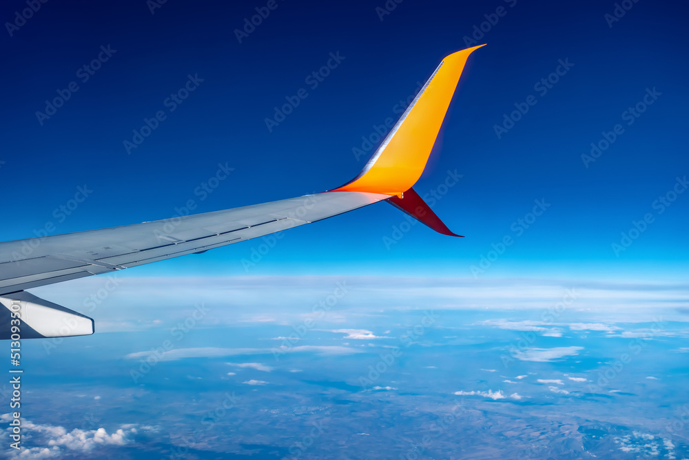 Fototapeta premium Airplane wing flying high in the atmosphere against the background of blue air. White-orange plane in the sky above the clouds and the planet