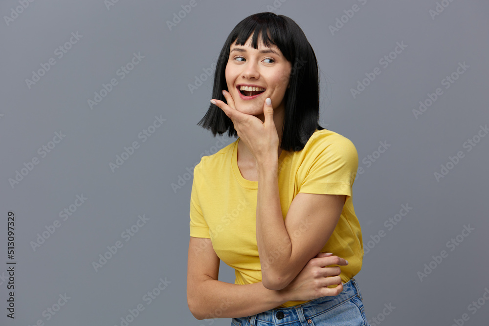 a sweet, pleasant, beautiful woman in a yellow T-shirt stands on a gray background, smiling pleasantly, holding her hand on her face