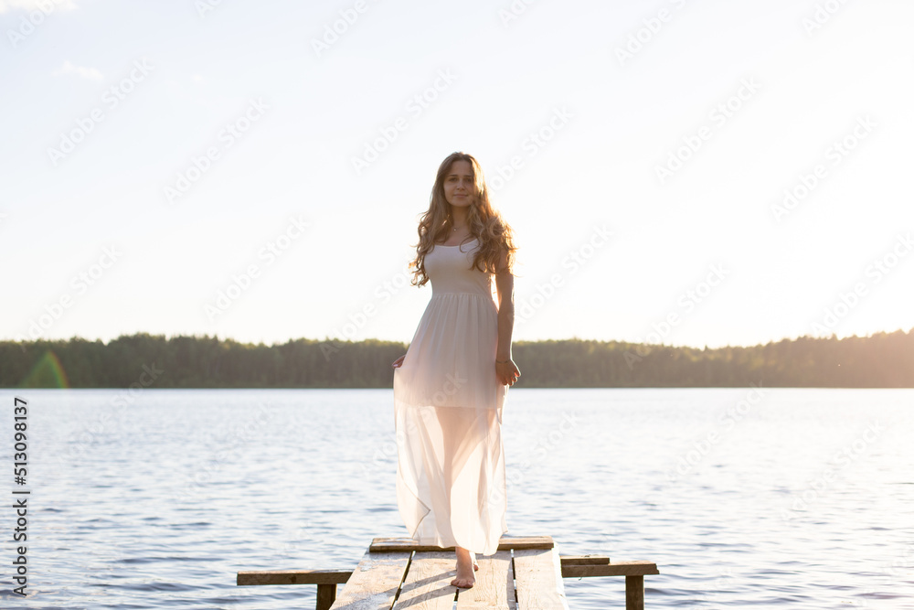 Beautiful Russian girl dressed in a white dress, walking along a wooden pier on the bank of a river or a lake