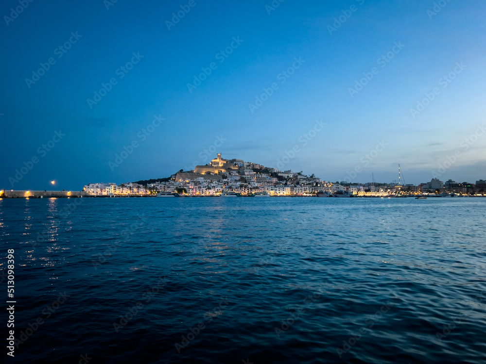 View of charming Ibiza Old town with it's castle, lit by evening lights, from the other side of the bay in Marina Botafoch. Enchanting view fora a romantic dinner for two. View of charming Ibiza Old town with it's castle, lit by evening lights, from the other side of the bay in Marina Botafoch. Enchanting view fora a romantic dinner for two.