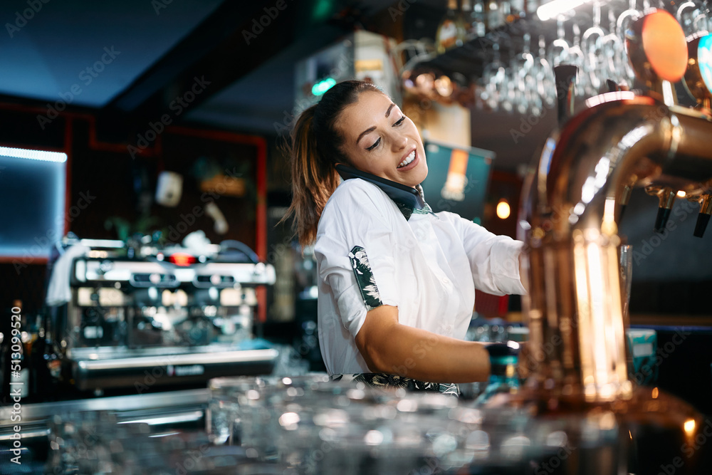 Young female bartender talks on phone while pouring beer from beer tap at bar counter. Young female bartender talks on phone while pouring beer from beer tap at bar counter.