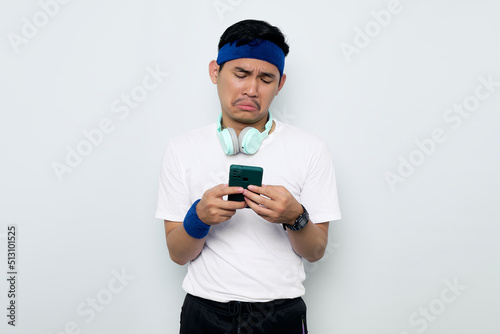 Disappointed young Asian sporty man in blue headband and white t-shirt with headphones, using mobile phone and reading bad news isolated on white background. Workout sport concept © Bangun Stock Photo