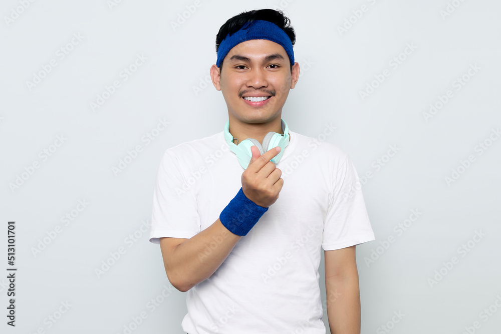 Portrait of smiling young Asian sportman in blue headband and white t-shirt with headphones, showing Korean heart with one fingers crossed isolated on white background. Workout sport concept Portrait of smiling young Asian sportman in blue headband and white t-shirt with headphones, showing Korean heart with one fingers crossed isolated on white background. Workout sport concept