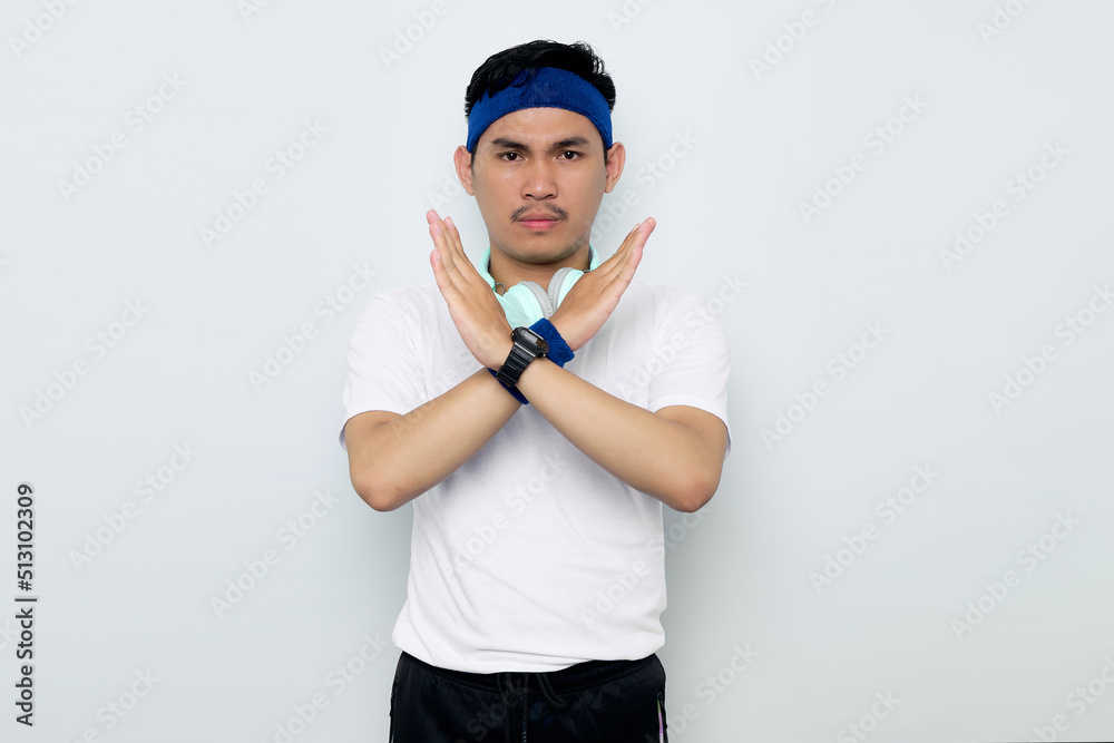 serious young Asian sportman in blue headband and sportswear white t-shirt with headphones,showing stop gesture, demonstrating denial sign, rejecting something unwanted isolated on white background serious young Asian sportman in blue headband and sportswear white t-shirt with headphones,showing stop gesture, demonstrating denial sign, rejecting something unwanted isolated on white background