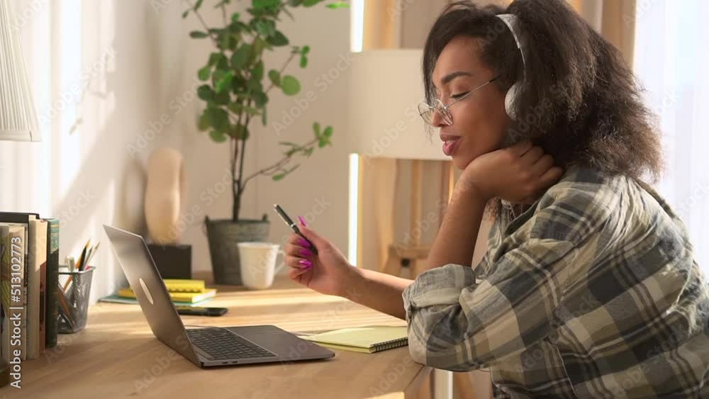 Young woman in headphones is studying online in front of laptop at table at home spbd. Close view of American African female looks at display with smile and listens to webinar, sits at desk in bright Young woman in headphones is studying online in front of laptop at table at home spbd. Close view of American African female looks at display with smile and listens to webinar, sits at desk in bright