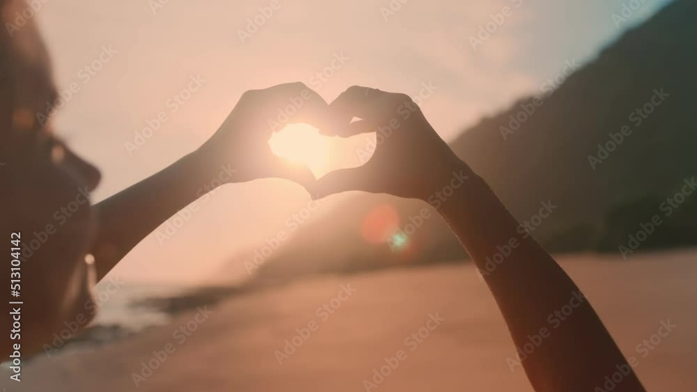 Young happy Caucasian woman teen with hair blowing in wind smiling making heart symbol with hands demonstrating love for this vacation spot hoping to return in future stands on beach near sea