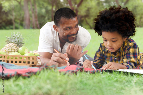 Happy smiling African American father and son go out for picnic outdoor in park on holiday, boy with curly hair drawing picture, lying on green grass with dad, joyful family playing outside in nature.