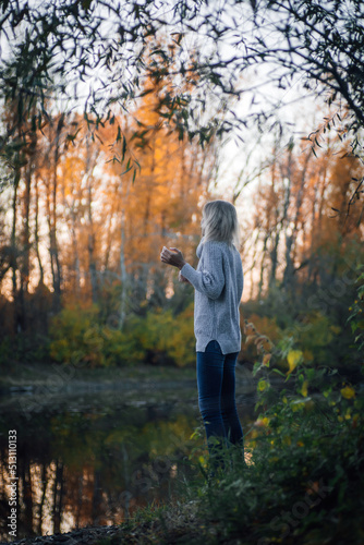 Peaceful blonde woman standing by river in forest, looking away at sunset and posing with trees in background being part of nature, wearing blue jeans and grey sweater. Autumn walk in forest. © Юля Бурмистрова
