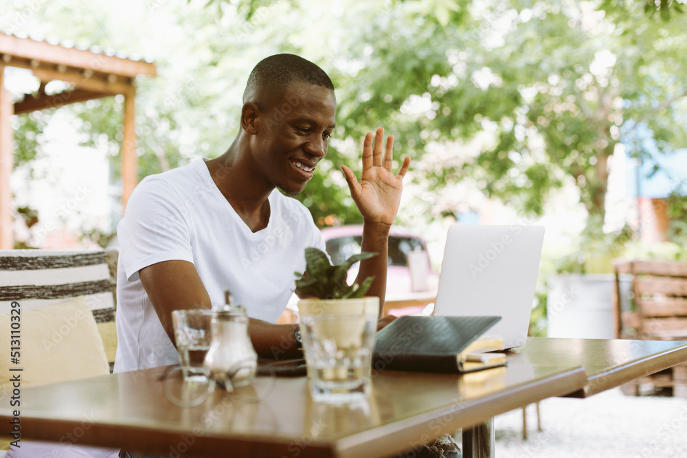 Smiling and glad dark skin man with laptop, raising arm, greeting someone, looking at monitor. E-learning webinar, video call, online meeting tutorial in coffee shop. Success work. Remote online job Smiling and glad dark skin man with laptop, raising arm, greeting someone, looking at monitor. E-learning webinar, video call, online meeting tutorial in coffee shop. Success work. Remote online job