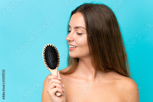 Young caucasian woman isolated on blue background with hair comb and looking it