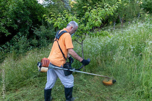 A mature man in protective clothing, and gloves with a trimmer. Mowing tall grass and the weeds with a trimmer. Selectively focus