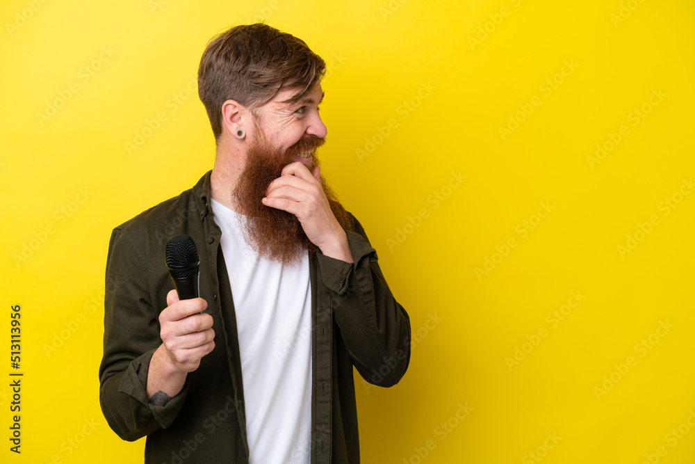 Redhead man with beard picking up a microphone isolated on yellow background thinking an idea and looking side Redhead man with beard picking up a microphone isolated on yellow background thinking an idea and looking side