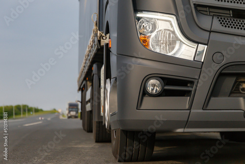 The concept of cargo transportation. A black truck is parked on the side of the highway.  Front view of a heavy truck. Front headlight close-up. A place to copy.