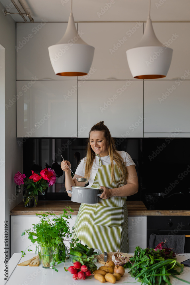 Vertical plump smiling blond woman in apron hold pan and ladle, cooking healthy vegetable soup in kitchen. Healthy diet