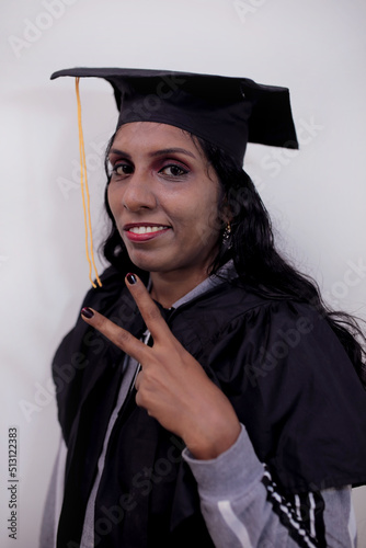 Young woman wearing graduation cap and ceremony robe smiling and confident gesturing with hand doing victory sign with fingers looking and the camera