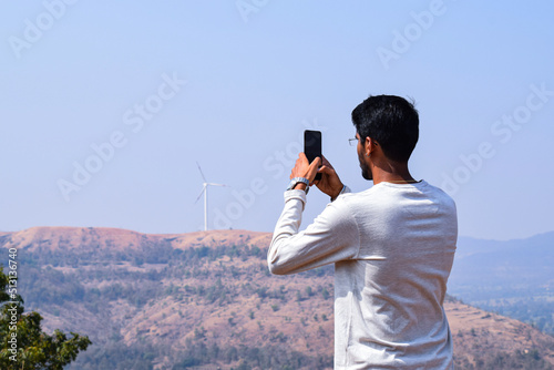  Back view of 25 to 30 age Indian man wearing white color t shirt and spectacles, clicking some photographs from his smart phone of the hills under bright sunlight. photo