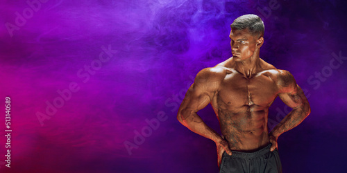 A handsome confident athletic man with bare mscular torso posing on a dark background filled with smoke. Male beauty concept. Fitness, bodybuilding