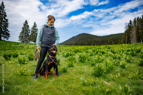 Teenage girl stands with leash in hands next to dog of Rottweiler breed in meadow  against background of fir trees
