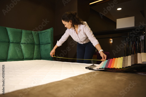 Pretty saleswoman holds a measure tape measuring the orthopedic mattress dimensions in a furniture design showroom. Upholstery fabric samples lying down on the soft bed